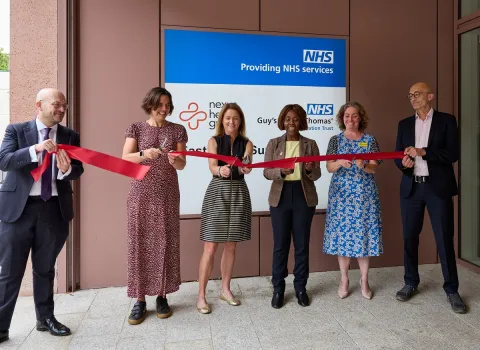 Six people standing in front of an NHS sign. They are holding a red ribbon and scissors