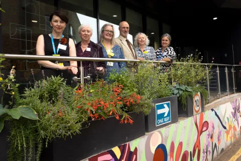 Group of six people standing behind a railing adorned with green plants and red flowers outside a building. They are overlooking a colourful mural.