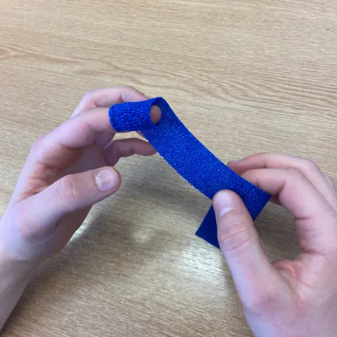 Photo of a pair of hands holding the ends of a long, thin dark blue bandage. One end of this bandage has been wrapped around the tip of the left index finger.