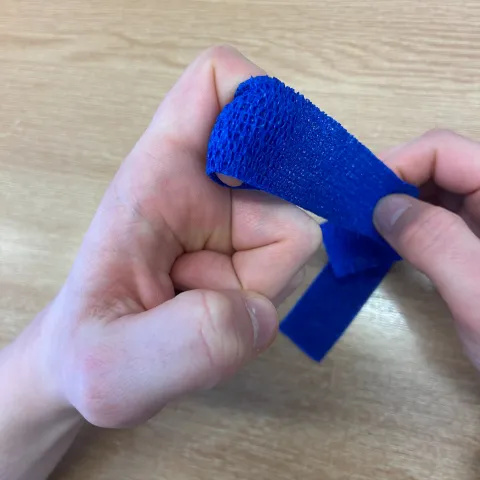 Photo of a pair of hands and a long, thin dark blue bandage. This bandage is being wrapped around a bent left index finger while the other hand is pulling the bandage to tighten it.