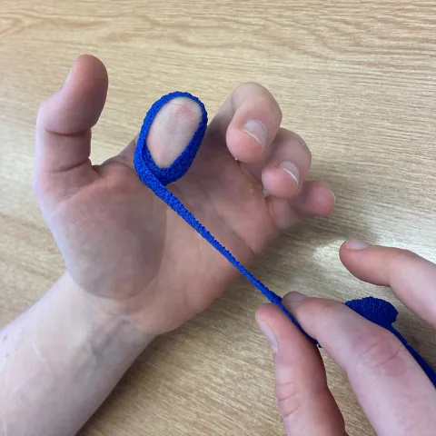 Photo of a long, thin dark blue bandage. One end is wrapped around a bent left index finger in a hook position and the right hand holds the other end.