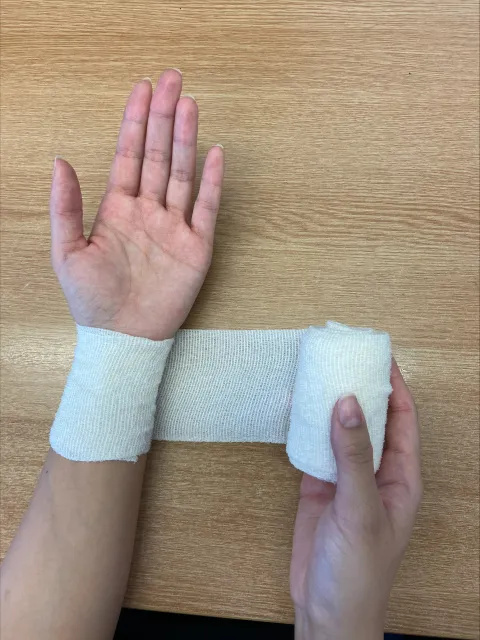Photo of someone's right hand holding a roll of white bandage. This has been wrapped around their left wrist. The palm and fingers of the left hand are outstretched.