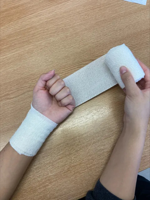 Photo of someone's hands and forearms. The right hand is holding a roll of white bandage, which is positioned behind the person's left hand. The bandage has been wrapped around the person's left wrist. The fingers on the person's left hand are bent to make a fist and pulled down towards the palm.