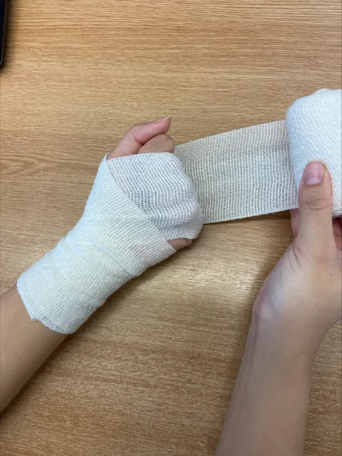 Photo of someone's hands and forearms. A white bandage has been wrapped around their left wrist and over the fingers on their left hand. The right hand is holding the roll of bandage, which is positioned behind the person's left hand.