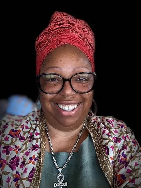 A woman wears a colourful red headscarf, a purple and white floral scarf, a green top, a necklace and black glasses