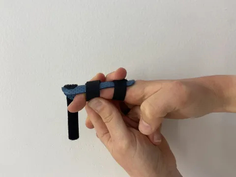 Photo of a pair of hands. There is a splint with 3 straps on the index finger of the person's right hand. The top strap is undone. The person is using their left thumb and hand to block the right index finger just below the crease of the tip. They are also starting to bend the tip of their right index finger.