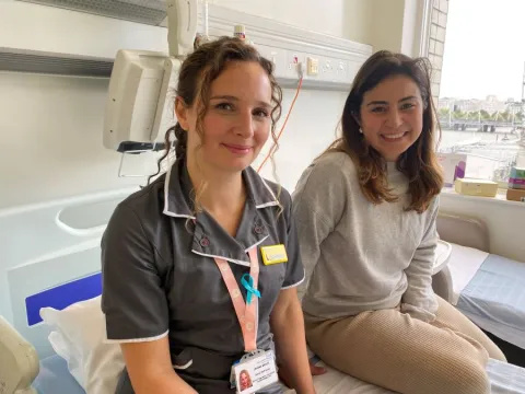 Nurse wearing a dark uniform with a name badge sits beside a woman in casual clothes on a hospital bed near a window overlooking London.