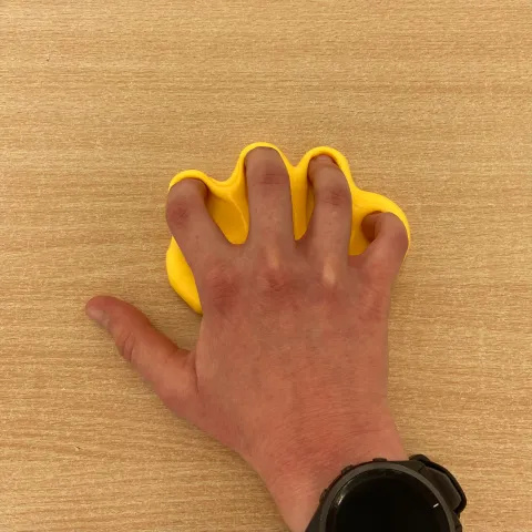 Photo of someone's hand with some flattened yellow putty nearby on a table. They are gripping the putty with closed fingers in preparation for flicking it.