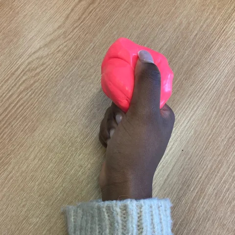 Photo of someone's hand with their fingers bent and the thumb straight. They are pinching some red putty between their thumb and index finger.