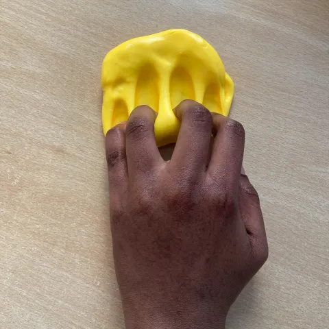 Photo of someone's hand with some flattened yellow putty nearby on a table. They are scratching the putty with bent fingers.