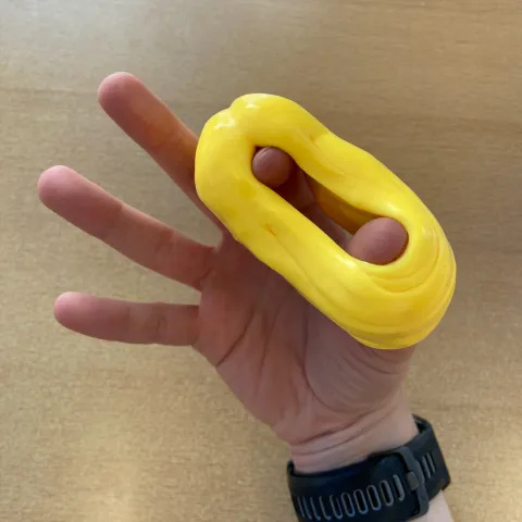 Photo of someone's hand with a large yellow ring of putty around their thumb and index finger. They have spread their thumb and index finger open widely.