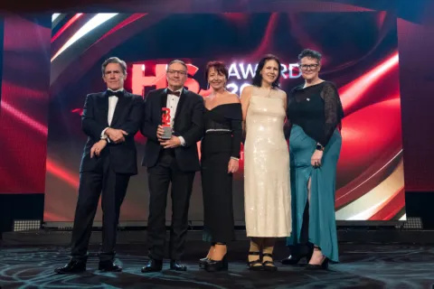 Five formally dressed individuals standing on stage at an awards event with a large illuminated backdrop behind them.