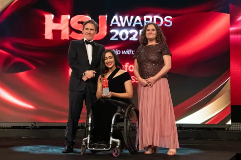 Three individuals pose on stage at the HSJ Awards 2025, with one person in a wheelchair holding a trophy, dressed formally.