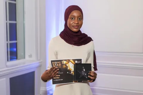 A woman wearing a white tunic and a burgundy headscarf is holding a certificate marked 'Rising Star' and an engraved object.