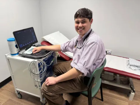 George is wearing a checked shirt and khaki cords. He is sitting at an echo machine, and there is a clinic bed behind him.