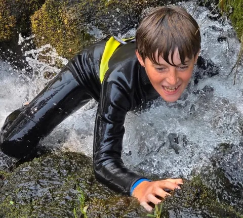 James is wearing a black wetsuit with a yellow flash. He is swimming in white water, across rocks.