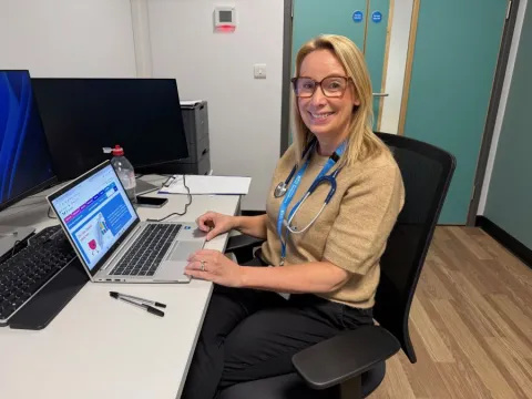 Leoni Bryan, valve nurse, is sitting at a desk and laptop in a clinic room. She has a stethoscope round her neck and is wearing a beige top.