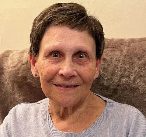 Patient Theresa Meli has short dark hair. She is sitting on a sof, wearing a blue top, and is smiling.