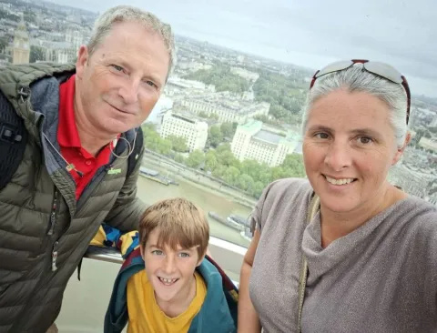 James is in a London Eye pod, wearing a yellow t-shirt and jacket. His dad Del has on a red t-shirt and dark green jacked, while mum Suzanne has a grey top on.