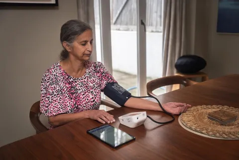 A woman sits at a table at home and takes a blood pressure reading