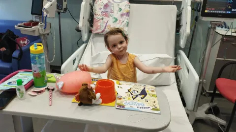 Luna in a yellow top in a hospital bed in front of her toys with her arms outstretched.