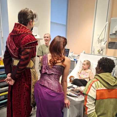 Sienna is sitting on a hospital bed and her mum is standing next to the bed, 3 of the Hercules cast members in costume are standing at the end of the bed