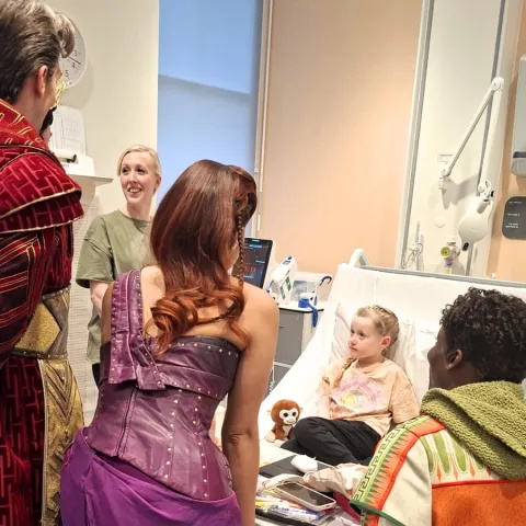 Sienna is sitting on a hospital bed and her mum is standing next to the bed, 3 of the Hercules cast members in costume are standing at the end of the bed
