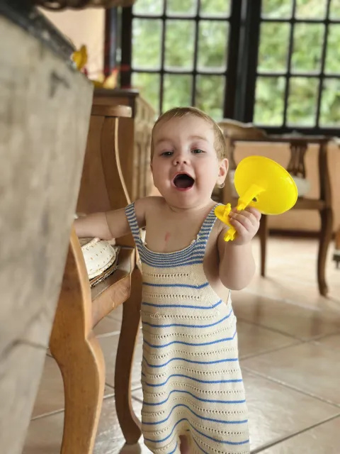 Atlas as a toddler, standing up in dungarees holding a yellow toy and leaning on a chair.