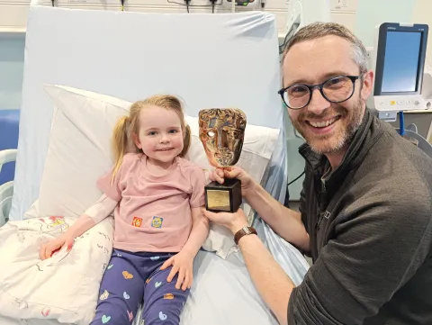 Luna in a hospital bed with Stuart next to her, both are smiling and Stuart is holding a Bafta award. 