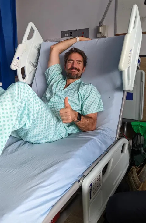 Chris lying in a hospital bed giving a thumbs up. 