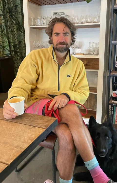 Chris wearing a yellow jumper and shorts drinking a coffee.