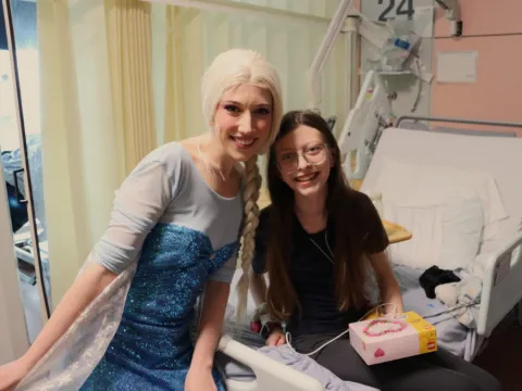 Sofia is sitting on a hospital bed, next to the bed is an entertainer dressed as the Disney princess Elsa, they're both smiling at the camera
