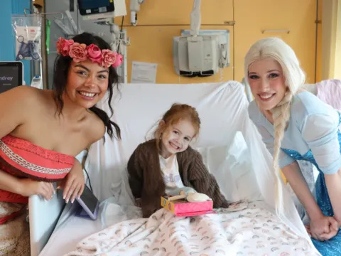 A child sitting on a hospital bed, standing next to the bed are two entertainers dressed as Disney princesses Moana and Elsa, they're all smiling at the camera