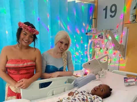 A child in a hospital bed, two entertainers are standing next to the bed, dressed as Disney princesses Moana and Elsa