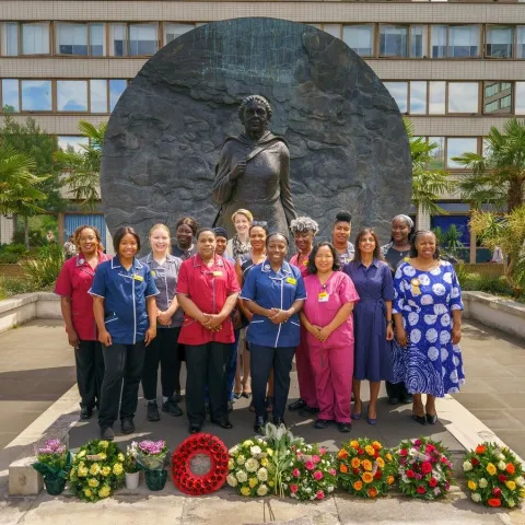 A large group of healthcare professionals and staff stand in front of a large bronze statue. Wreaths and flowers are in front of them.