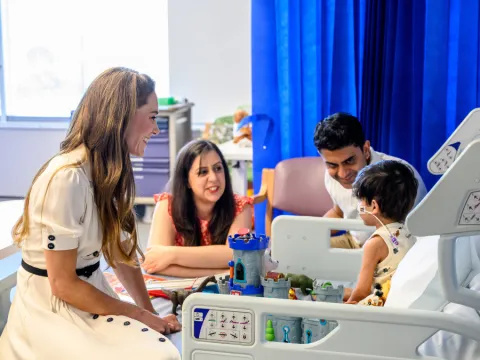 HRH The Princess of Wales sitting on a hospital bed, smiling at Arjun, who is on the bed. His mum and dad are sitting next to the bed, also smiling at Arjun. 