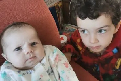Baby Alana in a white onesie on a red sofa next to her brother, who is wearing a red jumper. 