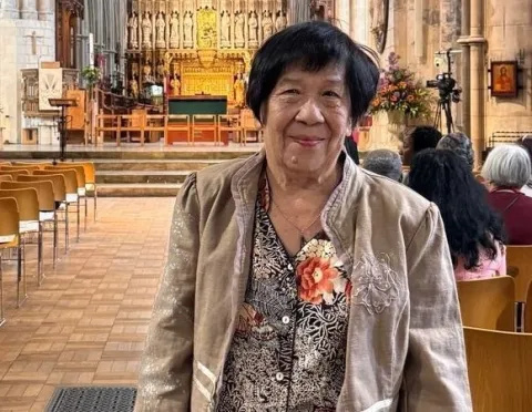 Christine Tan, wearing a beige jacket with a flower, is standing in a church