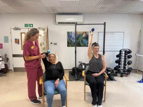 A physio, in maroon scrubs, is helping a seated patient to lift a dumbell. Another patient is seated nearby, and is lifting a dumbell above her head with her right arm.