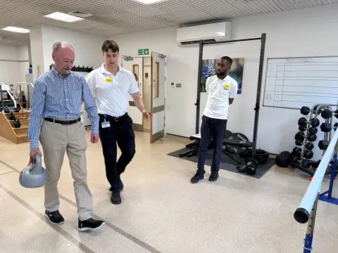 Patient Kevin, with physios James Cameron and Shak Springer in the hospital gym. Kevin is walking with a weight in his right hand, with James next to him.