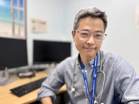 Dr Ronny Cheung, consultant paediatrician at Evelina London, sits turned away from his desk and computer and has a warm expression. He wears a grey shirt, blue lanyard and a stethoscope