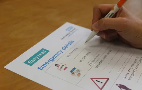 Alt text A hand holding a pan is completing a paper form. The form says 'Easy read' at the top of the page, and contains blank spaces to complete about a patient's emergency details.
