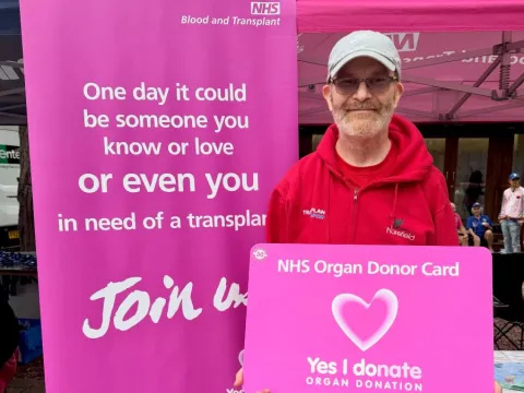 Dave is wearing a red hoodie, and is holding a giant pink Organ Donation card. he is standing next to a pink banner also promoting organ donation, from NHSBT.