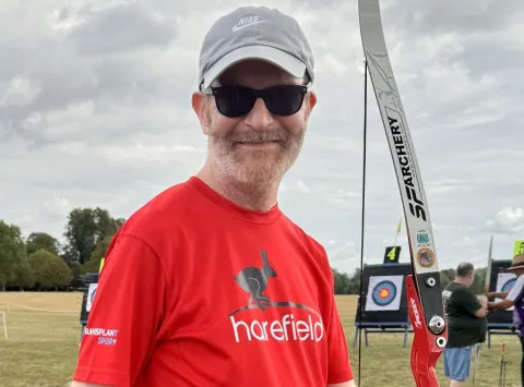 Dave is wearing a red t-shirt with Harefield written in white on the front. He is holding an archery bow, and is standing in front of targets.