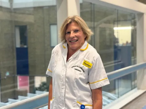 Carolyn Doughty is wearing a white uniform tunic with yellow trim. She is standing in a corridor with glass panels behind her, in Guy's Hospital