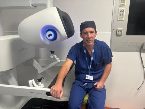 Professor James Gossage, dressed in blue surgical scrubs, is sitting in front of the console of the da Vinci 5 robotic-assisted surgical system, which has been fitted with the new Force Feedback technology.