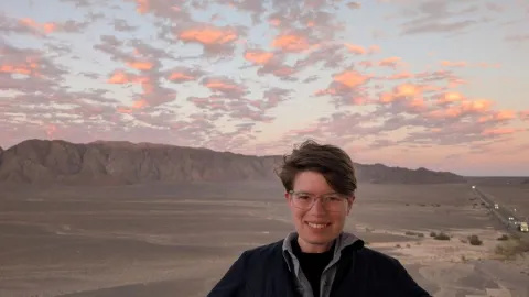 Imogen is standing in front of a desert, with a pink cloudy sky in the background.