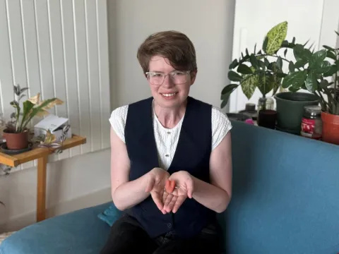 Imogen, wearing a white top and navy waistcoat, is sitting on a sofa, holding her prosthetic tongue in her hands in front of her.