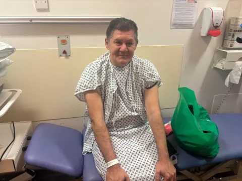 Patient Nicholas Kokkinos is pictured sitting on a blue clinical bed ahead of his surgery. He is smiling and wearing a hospital gown and a hospital ID bracelet.