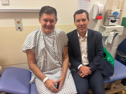 Patient Nicholas Kokkinos and surgeon Professor James Gossage are pictured sitting next to each other on a blue clinical bed. Nicholas is wearing a hospital gown and James is wearing a white shirt and dark suit.